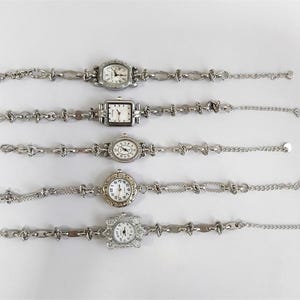 May include: Five silver-toned wristwatches with various face shapes: oval, square, and round. Each watch has a different band design, including chain and link styles. The watch faces are white with black numerals and hands.