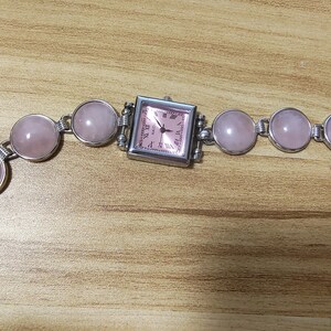 May include: A silver-toned bracelet watch with a square pink watch face and a series of round, pink gemstones. The watch face has Roman numerals and hands. The bracelet is made of silver links.