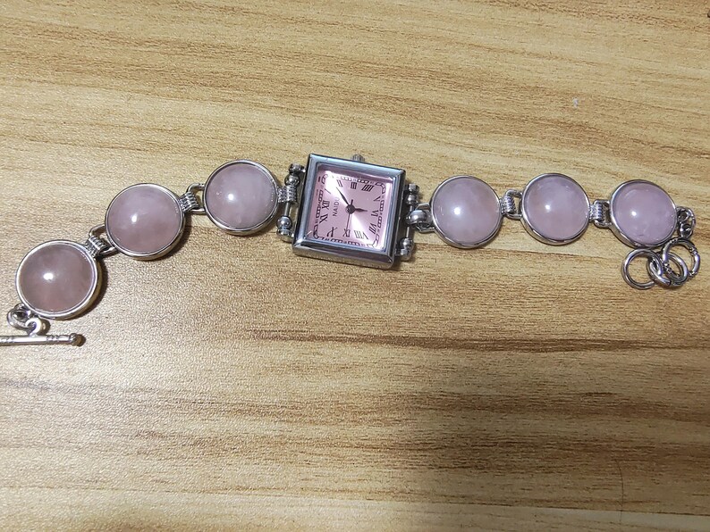 May include: A silver-toned bracelet watch with a square pink watch face and a series of round, pink gemstones. The watch face has Roman numerals and hands. The bracelet is made of silver links.