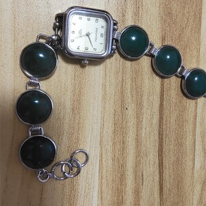 May include: A silver-toned wristwatch with a square face and a cream-colored dial. The watch band features large, round, dark green stones connected by silver links. The watch face displays the time with black numerals.