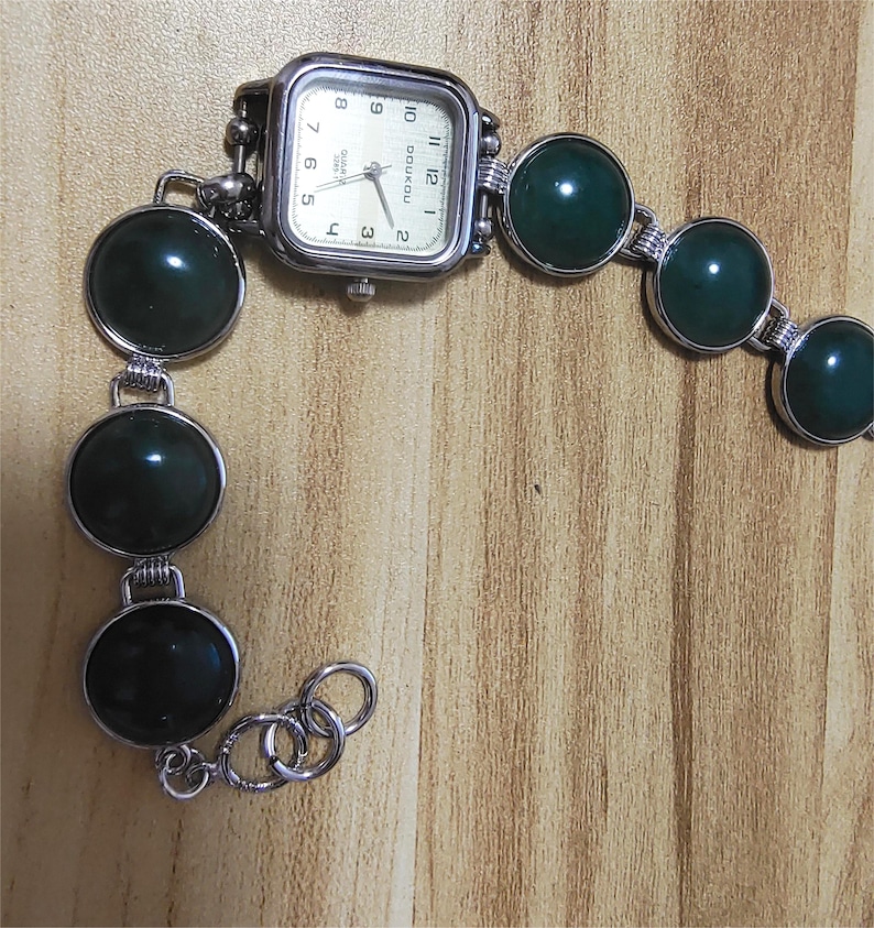 May include: A silver-toned wristwatch with a square face and a cream-colored dial. The watch band features large, round, dark green stones connected by silver links. The watch face displays the time with black numerals.