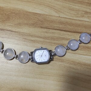May include: A silver-toned watch bracelet with a white watch face and a series of large, round, light-colored stones. The watch face is octagonal. The stones are connected by small silver links.