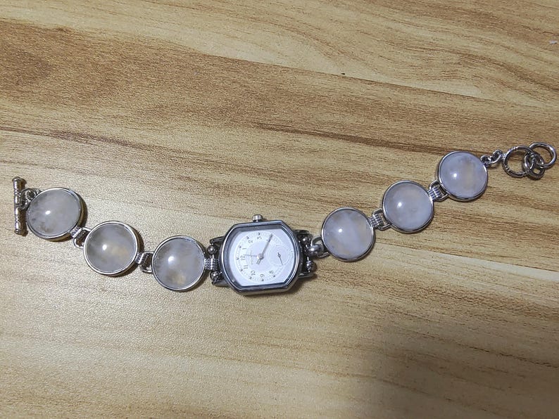 May include: A silver-toned watch bracelet with a white watch face and a series of large, round, light-colored stones. The watch face is octagonal. The stones are connected by small silver links.