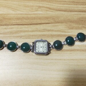 May include: A silver-toned watch with a square face and a cream-colored dial. The watch band features large, round, dark green stones. The watch is displayed on a light brown wooden surface.