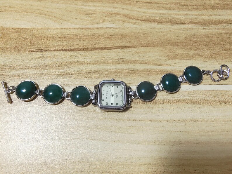 May include: A silver-toned watch with a square face and a cream-colored dial. The watch band features large, round, dark green stones. The watch is displayed on a light brown wooden surface.