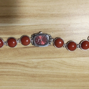 May include: A silver-toned wristwatch with a rectangular red face and a band featuring large, round, reddish-brown stones. The watch is set against a light brown wooden surface.