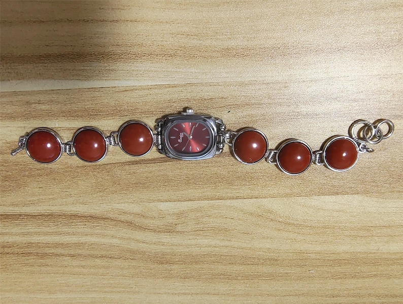 May include: A silver-toned wristwatch with a rectangular red face and a band featuring large, round, reddish-brown stones. The watch is set against a light brown wooden surface.