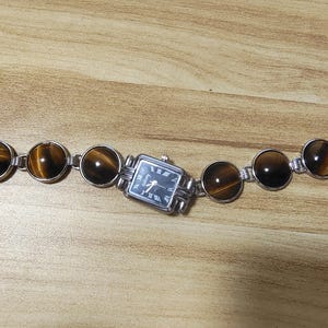 May include: A silver-toned bracelet watch featuring a rectangular watch face with black Roman numerals and hands. The bracelet is adorned with round, polished tiger's eye stones and connected by silver links. The watch is displayed on a wooden surface.
