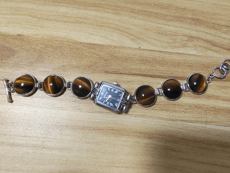 May include: A silver-toned bracelet watch featuring a rectangular watch face with black Roman numerals and hands. The bracelet is adorned with round, polished tiger's eye stones and connected by silver links. The watch is displayed on a wooden surface.