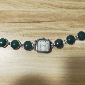 May include: Silver-toned watch with a square face and a series of dark green, circular stones. The watch band features a toggle clasp. The watch face has a white background with black numerals and hands.