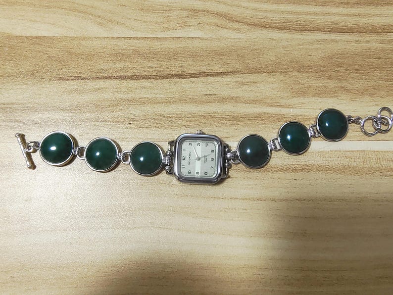 May include: Silver-toned watch with a square face and a series of dark green, circular stones. The watch band features a toggle clasp. The watch face has a white background with black numerals and hands.