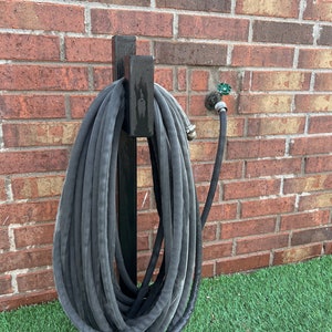 May include: A black metal hose holder mounted on a brick wall with a black garden hose wrapped around it. The hose holder has a hook on the top for hanging the hose.