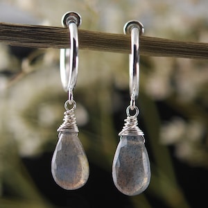 May include: A pair of silver hoop earrings with teardrop-shaped labradorite stones hanging from them. The stones are a dark grey colour with iridescent flashes of blue and green.
