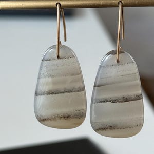 May include: A pair of teardrop-shaped earrings. Each earring features a translucent, light-colored stone with horizontal bands of gray and brown. The earrings are suspended from gold-colored, curved ear wires, creating a minimalist and elegant design.