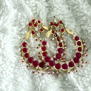 Puede incluir: Pendientes de aro dorados con diseño floral y gemas rojas. Los pendientes están adornados con cristales transparentes y pequeñas perlas blancas. Los aros tienen aproximadamente 5 cm de diámetro.