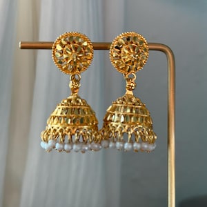 May include: A pair of gold-colored jhumka earrings. The earrings feature a circular top with a floral design, connected to a bell-shaped bottom. The bottom of the earrings are adorned with small white pearls. The earrings are displayed on a gold-colored stand.