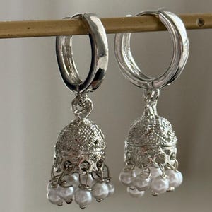 May include: Silver hoop earrings with a dangling charm featuring a silver bell design and white pearl accents.