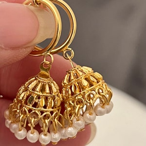May include: A pair of gold hoop earrings with dangling charms. The charms are shaped like bells and have small white beads hanging from them.