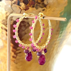 May include: A pair of gold hoop earrings featuring small, round, bright pink gemstone beads wrapped around the wire. Each hoop includes a dangling teardrop-shaped dark pink gemstone charm and a smaller round purple gemstone bead at the base.