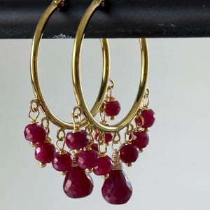 May include: A pair of gold hoop earrings featuring multiple dangling faceted deep pink gemstone beads. Each hoop is adorned with several small round beads and a larger teardrop-shaped bead at the centre, all suspended by thin gold wire links.
