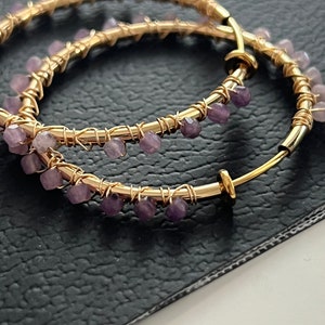 May include: Gold hoop earrings with purple gemstone accents. The earrings are made of wire wrapped around a hoop shape, with small purple gemstones attached to the wire.
