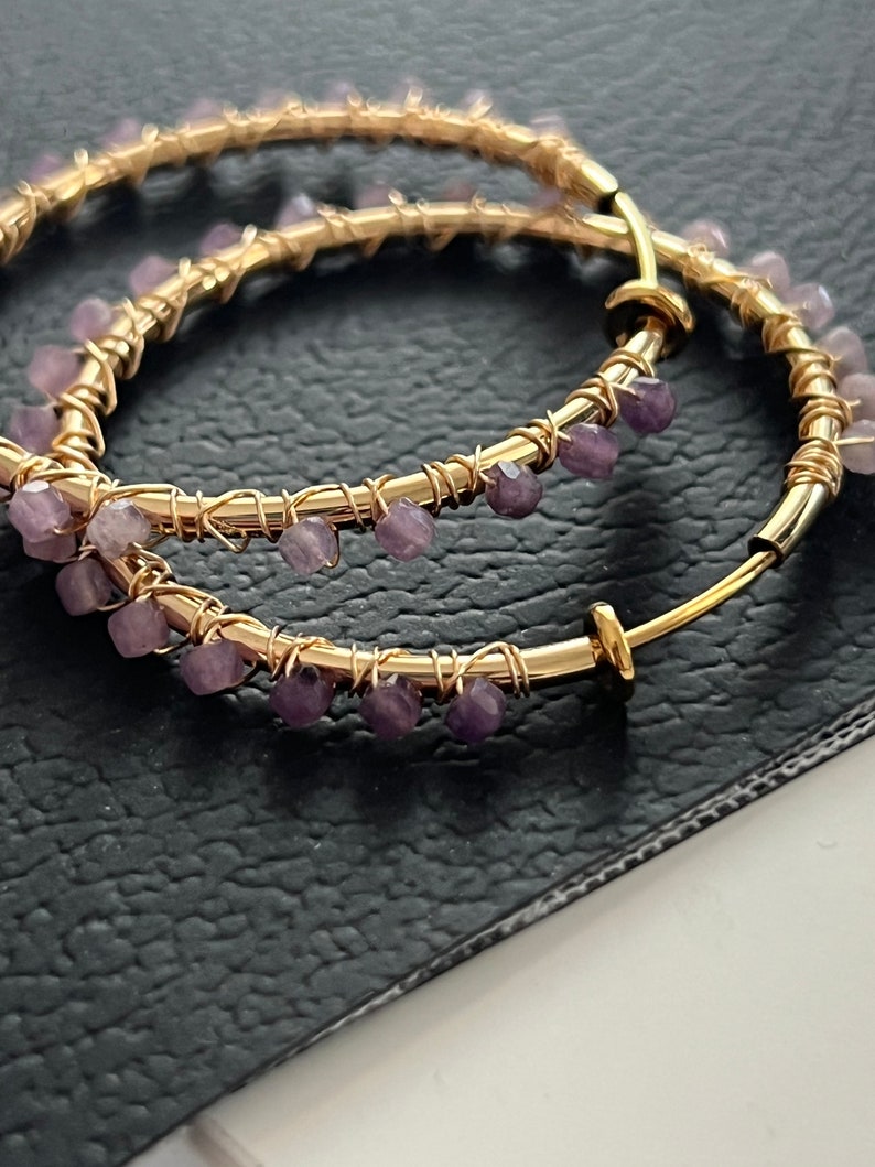 May include: Gold hoop earrings with purple gemstone accents. The earrings are made of wire wrapped around a hoop shape, with small purple gemstones attached to the wire.