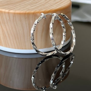 May include: A pair of silver hoop earrings with a twisted design. The earrings are on a wooden surface.