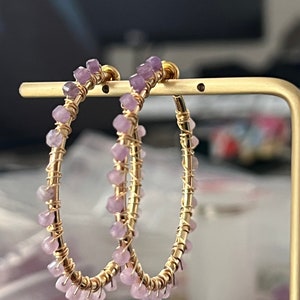 May include: A pair of gold hoop earrings with small purple gemstones. The earrings are displayed on a gold stand.
