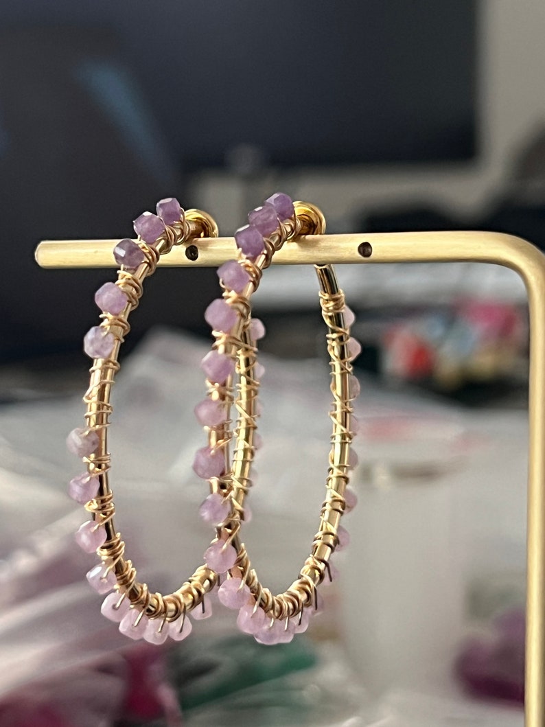 May include: A pair of gold hoop earrings with small purple gemstones. The earrings are displayed on a gold stand.