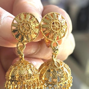 May include: A pair of gold-colored earrings. Each earring features a circular top with a floral design and a bell-shaped bottom. The earrings are connected by a delicate chain and adorned with small, dangling beads.