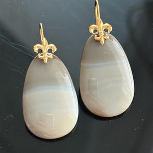 May include: A pair of teardrop-shaped earrings. Each earring features a polished, gray stone with a band of white and a gold-colored fleur-de-lis accent at the top. The earrings have a simple hook closure.