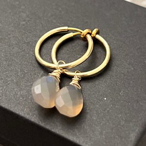 May include: Gold hoop earrings with a light gray gemstone dangling from each hoop.
