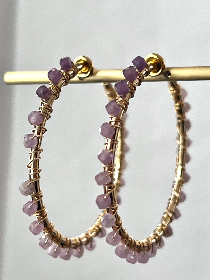 May include: Gold hoop earrings with a delicate wire design and small purple gemstones. The earrings are approximately 2 inches in diameter.