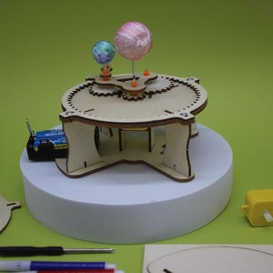 Könnte beinhalten: Ein Holzmodell eines Planetensystems mit zwei bunten Kugeln, die Planeten darstellen. Das Modell steht auf einer weißen Plattform vor einem grünen Hintergrund. Zusätzliche Komponenten, darunter eine Batterie und ein Motor, sind sichtbar.