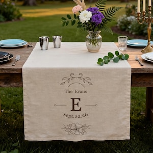 May include: A rustic wooden table set for an outdoor event. A beige linen table runner is centered, featuring the text "The Evans", the letter "E", and the date "sept.22.26". The table is decorated with flowers, candles, plates, and silverware.