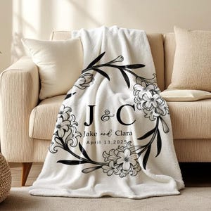 May include: A white fleece blanket draped over a beige sofa. The blanket features a black floral wreath design with the initials "J & C", the names "Jake and Clara", and the date "April 13.2025".