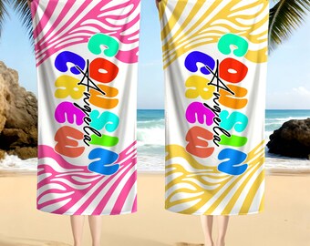 May include: Two beach towels with a white center and colorful text that reads "Corsica Angela". One towel has a pink zebra stripe pattern, the other has a yellow zebra stripe pattern. The towels are held up on a beach.