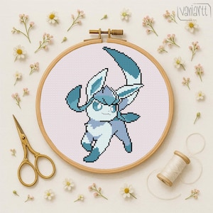 Puede incluir: Un bastidor de bordado de punto de cruz con un personaje de dibujos animados pixelado en tonos azules y blancos. El bordado está sobre una tela rosa claro, rodeado por un aro de madera. Tijeras doradas, hilo y flores completan la imagen temática de artesanía.