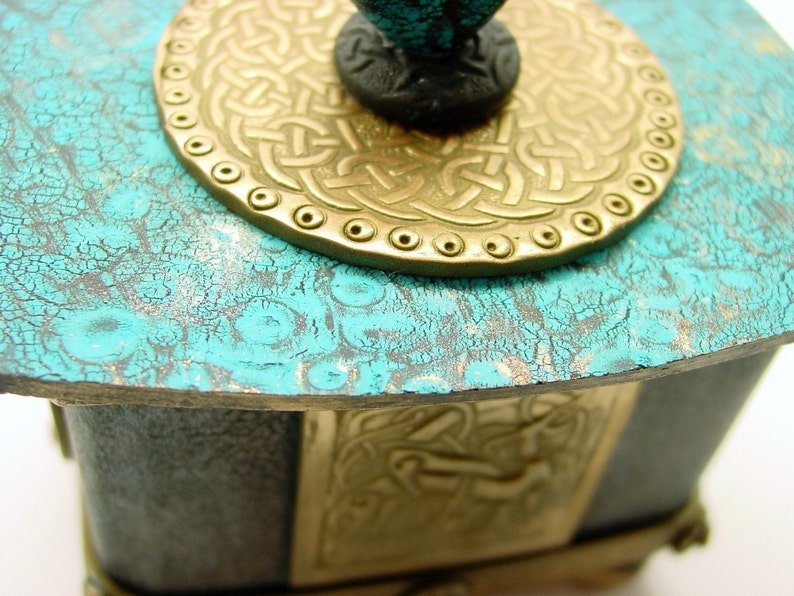 May include: A teal blue wooden box with a gold metal Celtic knot design lid. The lid has a raised, circular design with a black knob.