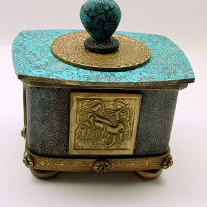 May include: A small, square, teal and gold decorative box with a turquoise knob. The box has a Celtic knot design on the front and a gold trim around the top and bottom.