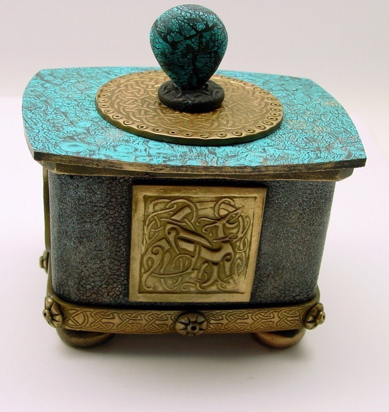 May include: A small, square, teal and gold decorative box with a turquoise knob. The box has a Celtic knot design on the front and a gold trim around the top and bottom.
