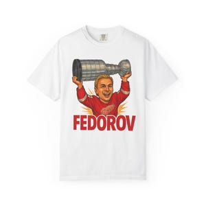 May include: White t-shirt featuring a cartoon illustration of a hockey player holding a trophy above his head. The player is wearing a red jersey with the name "FEDOROV" in red block letters below the illustration.