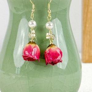 May include: A pair of gold earrings with a pink dried rose and a pearl bead on each earring.