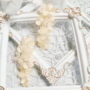 Puede incluir: Dos pinzas para el pelo florales blancas con un delicado diseño en cascada. Las pinzas se muestran sobre un fondo blanco con una superposición de encaje.