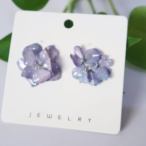 May include: A pair of purple flower earrings with iridescent accents. The earrings are displayed on a white card with the word "JEWELRY" printed on it.