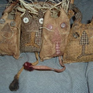 May include: Four handmade cat-shaped fabric dolls with button eyes and stitched details. The dolls are in various shades of brown and tan, with some featuring checkered patterns and tree-like designs. Each doll has a tail and is arranged in front of a straw nest.