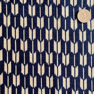 Può includere: Primo piano di un tessuto con un motivo ripetuto di forme a forma di freccia color crema su sfondo blu navy. Una moneta da un quarto di dollaro USA si trova nell'angolo in alto a destra per la scala. Il tessuto ha un aspetto strutturato.