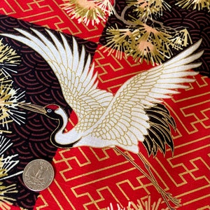 以下が含まれることがあります： 白い鶴のデザインと金のアクセントが施された赤い生地。鶴は翼を広げて飛んでいる。生地は金の正方形と白黒の円が繰り返されるパターンになっている。