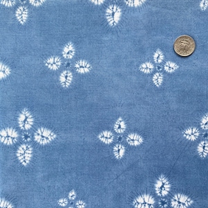 Può includere: Tessuto blu con un motivo ripetuto di forme floreali bianche a quattro petali. Il tessuto ha un aspetto strutturato e una moneta è visibile per la scala. Il design ricorda il tie-dye.