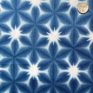 May include: A close-up of a fabric with a repeating floral pattern. The design features blue and white starburst-like flowers. The fabric has a textured appearance, with the blue dye creating a tie-dye effect. A coin is in the upper right corner.